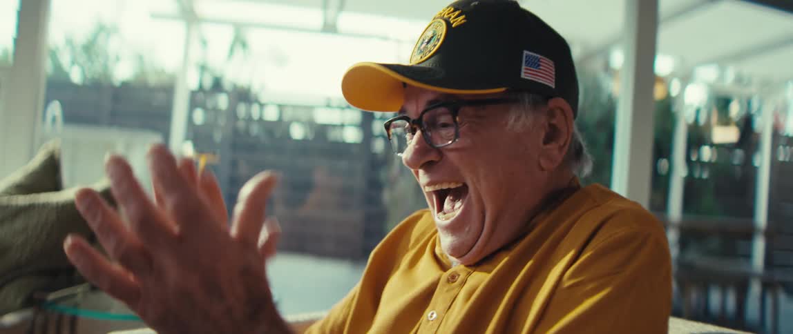 Elderly man wearing a cap and glasses sitting in an armchair laughing and clapping