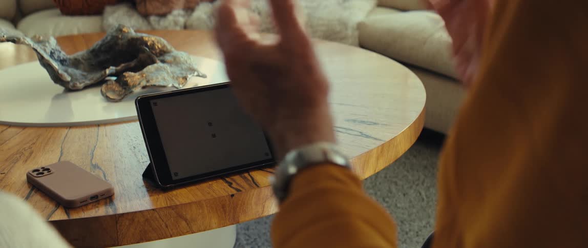 An older man wearing a yellow shirt and silver watch gestures with his hands while looking to a tablet resting on a round wooden coffee table next to a glass of water and a smartphone