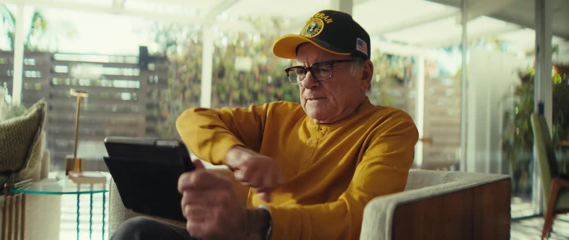 Elderly man wearing a cap and glasses sitting in an armchair looking at a tablet with his hand near his chin