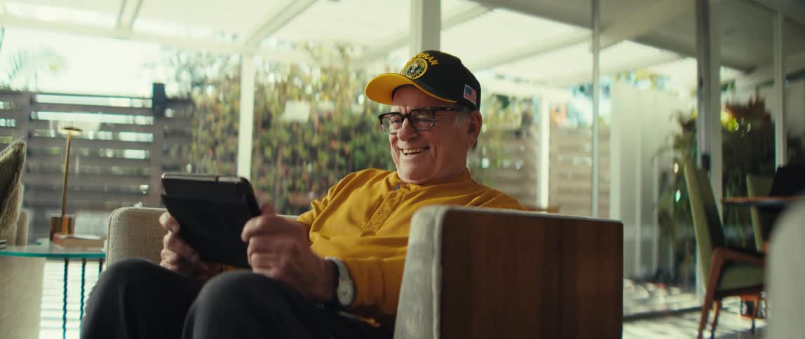 Elderly man wearing glasses and a baseball cap sitting on an armchair holding a tablet computer and laughing while talking indoors during day time