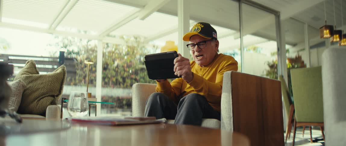 An older man wearing glasses and a veteran hat celebrates with excitement while holding a tablet in a bright living room