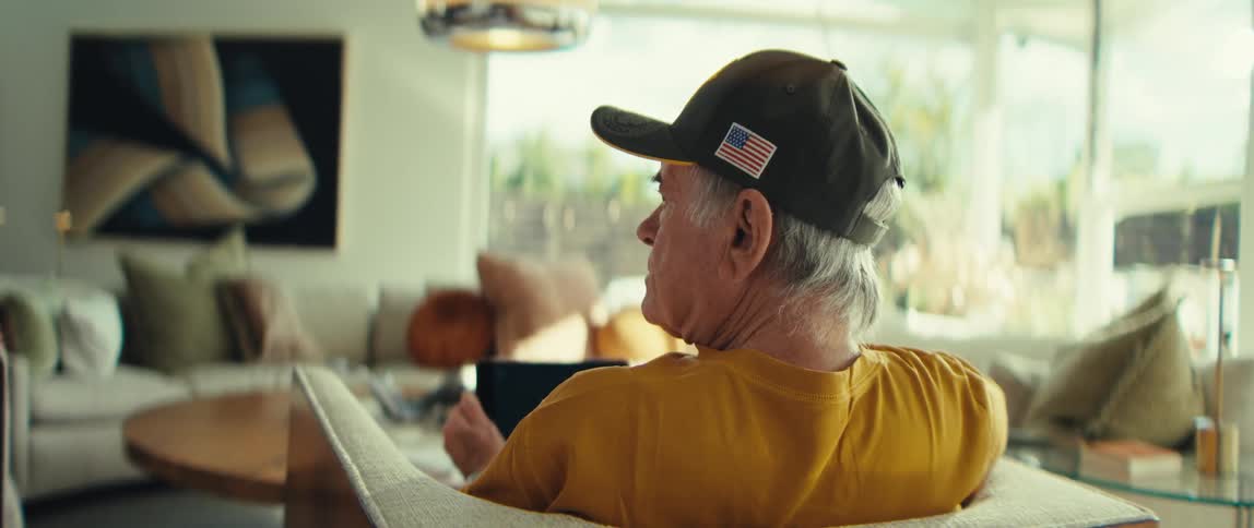 An older man with grey hair wearing a yellow shirt and a black cap sits on a white sofa in a bright modern living room while holding a tablet and looking to the left during the day