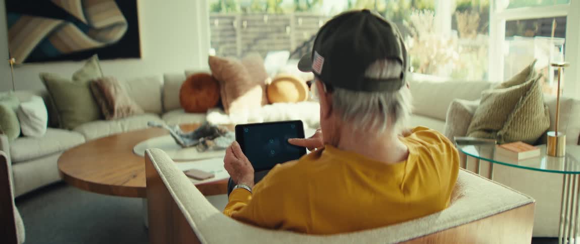 An older man wearing a cap sits on a couch in a bright living room holding and looking at a tablet