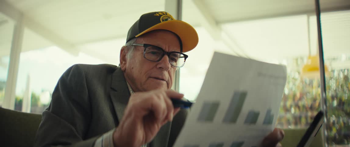 Elderly man wearing a blazer and a baseball cap sitting down on a couch looking and pointing with a pen at a paper with graphics on it indoors during day time
