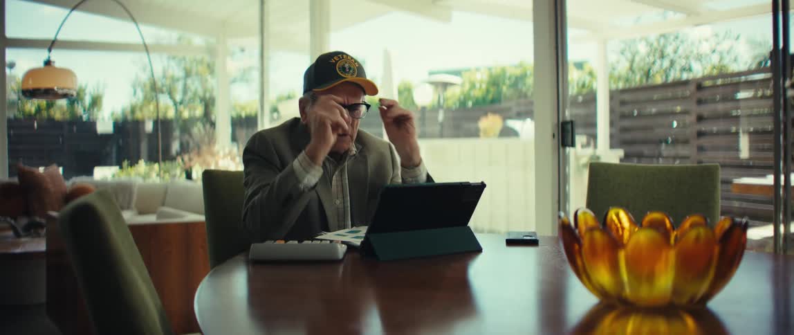 An elderly veteran wearing a baseball cap sits at a wooden table thoughtfully holding his glasses before putting them onhe then begins to interact with a tablet computer propped up in front of him which sits alongside a calculator and some paperwork