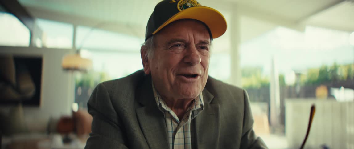 Elderly man wearing a cap and blazer laughing in a welllit indoor setting with glass doors in the background