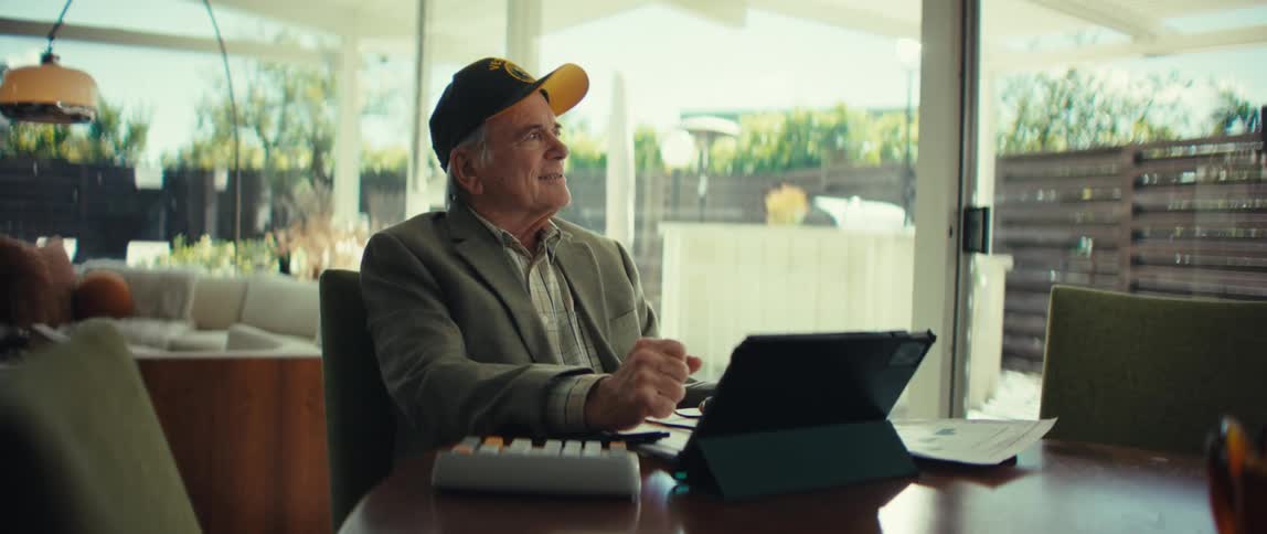 Man sitting at a table with a laptop computer looking at paperwork as he pauses to smile and think