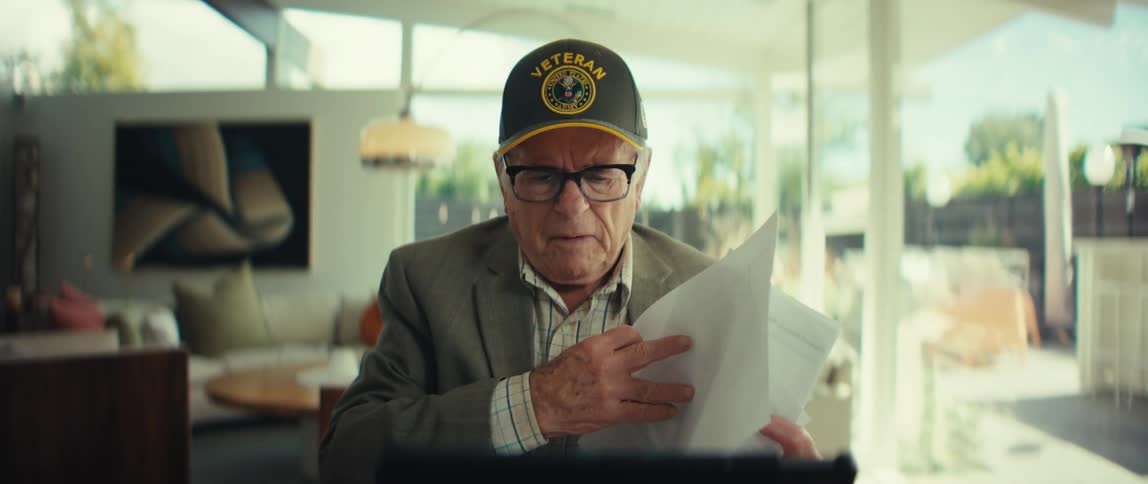Elderly man wearing a blazer and a cap with the word veteran written on it reading through papers with a nervous facial expression in a living room indoors during day time