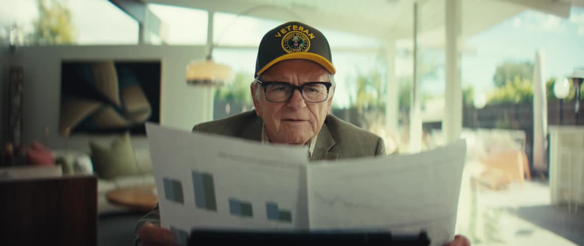 Elderly man wearing a cap and glasses holding a report with charts while looking at the document in a room with large windows in the background