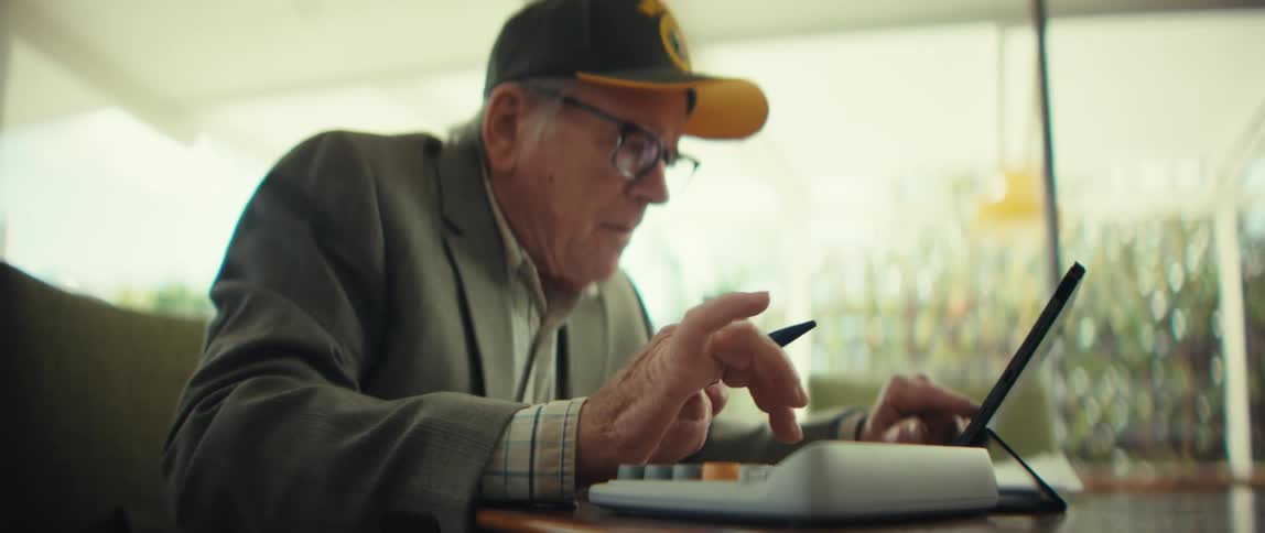 An elderly man wearing a yellow veteran hat and glasses sits at a sunlit table while focused on pressing buttons on a white calculator and holding a stylus near a tablet in an indoor setting