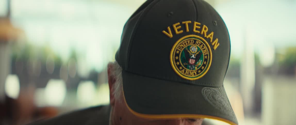 An elderly man sitting indoors at midday wearing a hat that says veteran on the front