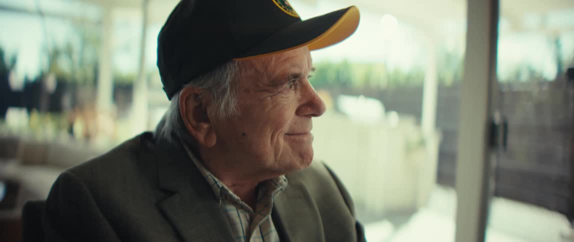 An elderly veteran wearing a black baseball cap and a gray suit jacket looks toward a bright window while smiling slightly in a soft natural light setting