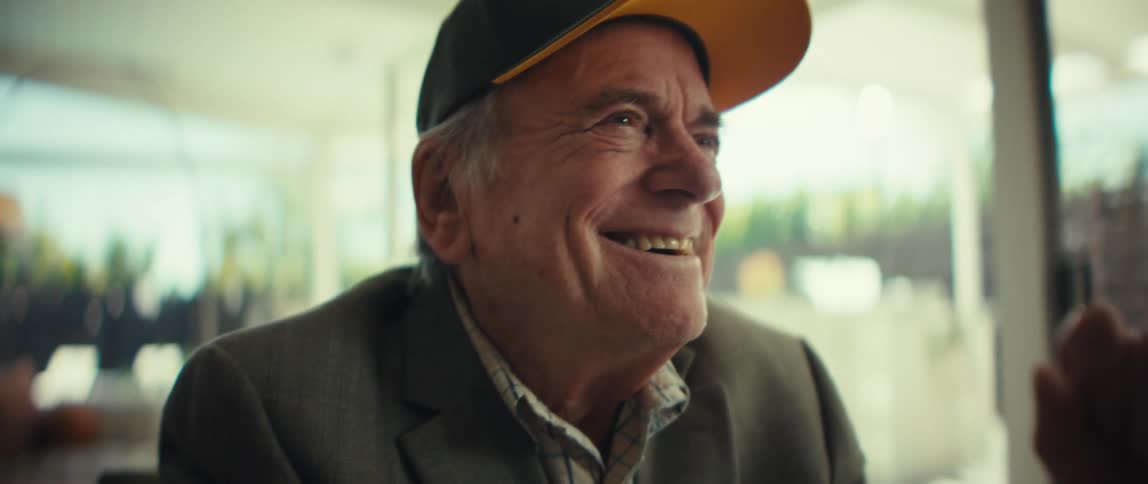 Elderly white man wearing a blazer and a baseball cap smiling and laughing while sitting indoors during day time