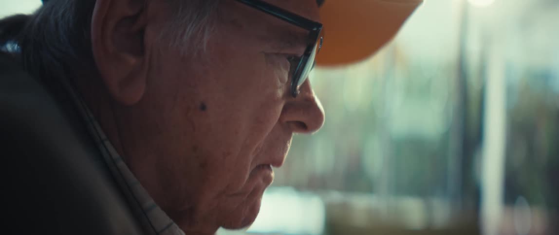 Elderly man wearing glasses and a cap with a focused expression in a welllit indoor setting