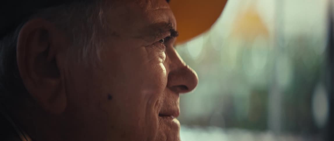 In a warm tight closeup an elderly man wearing a yellowbrimmed cap looks gently forward a soft and peaceful smile forming on his lined face as natural light catches his eyes