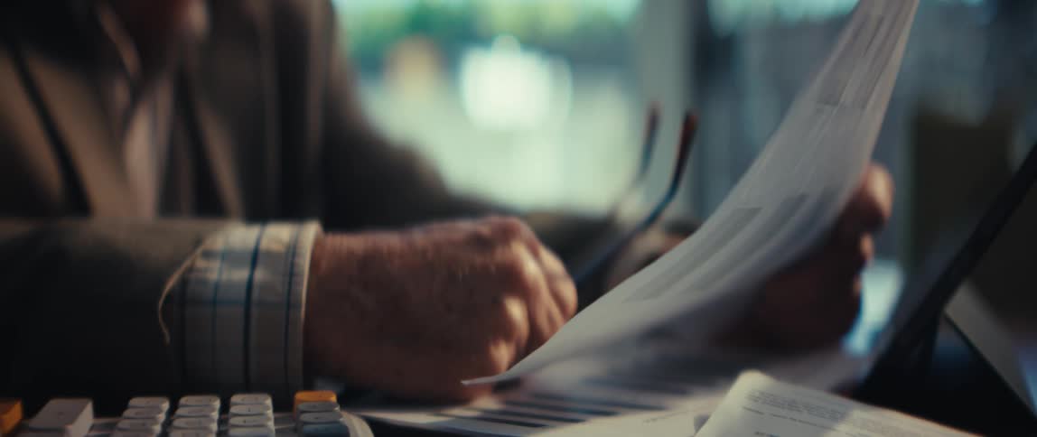 A close view of an elderly mans hands shows him holding eyeglasses while reviewing financial documents with bar charts at a desk in a dimly lit room