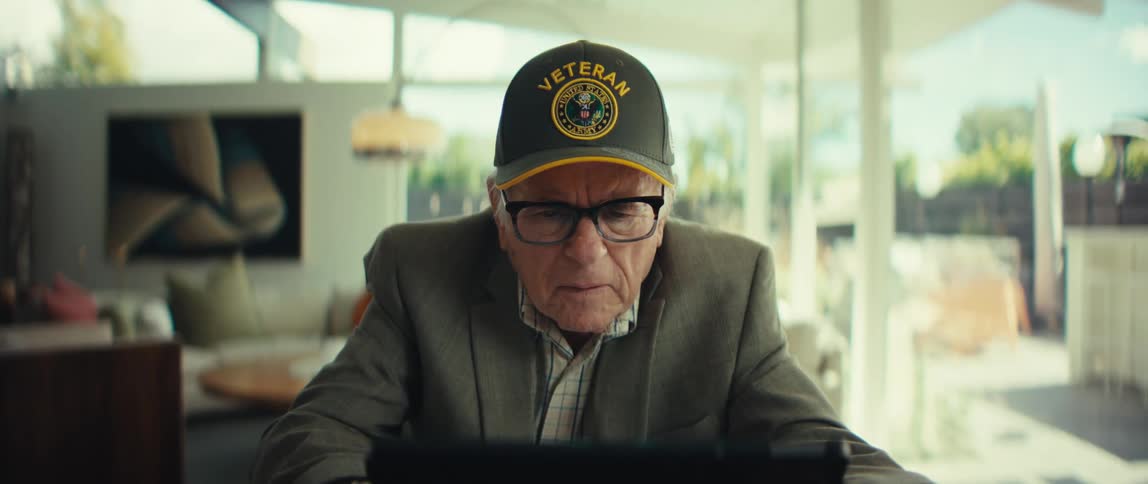Elderly man wearing a cap and glasses sitting at a table looking at a tablet in a room with large windows in the background