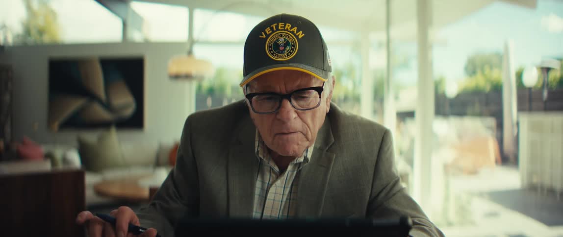 Elderly man wearing a blazer and a baseball cap sitting in front of a tablet computer with a nervous facial expression indoors during day time