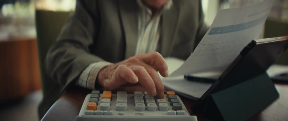 A man in a blazer sits at a desk and uses his finger to press buttons on a large white calculator while reviewing several paper documents and a tablet