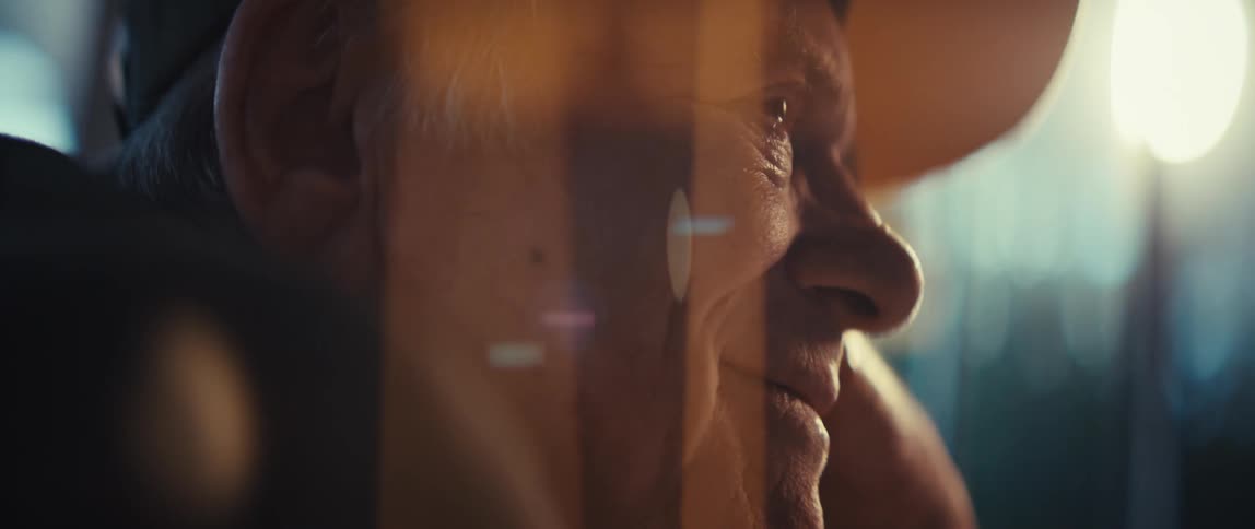 The video features an extreme closeup of an elderly man smiling warmly as he looks toward a soft light source overlaid reflections of glass and light flares drift across his face as he adjusts his glasses