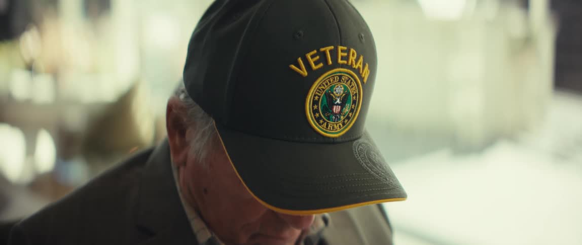 The video shows a closeup of an elderly man wearing a black baseball cap with the word veteran and a united states army seal embroidered on ithe is looking downward while wearing a blazer over a plaid shirt with the camera focus remaining tight on th
