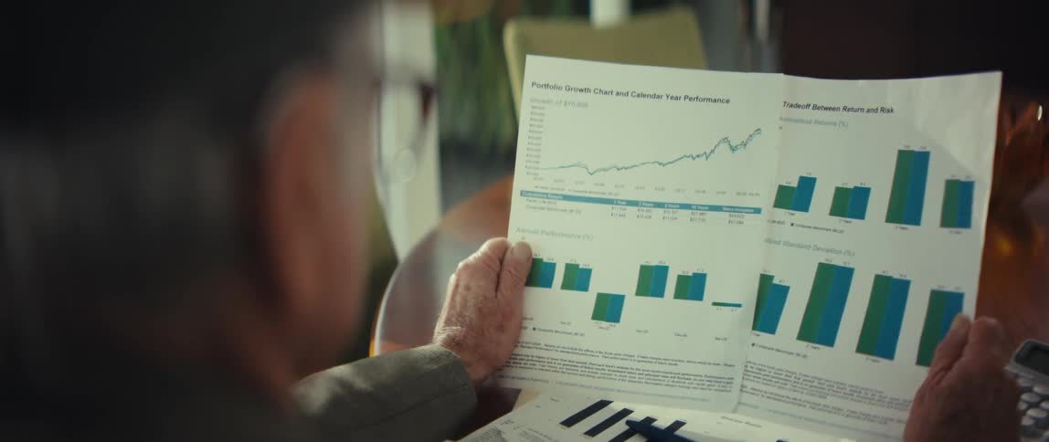 A view from behind an elderly man wearing glasses shows him holding a portfolio growth chart with several blue and green bar graphs and a line graph while sitting in a bright room