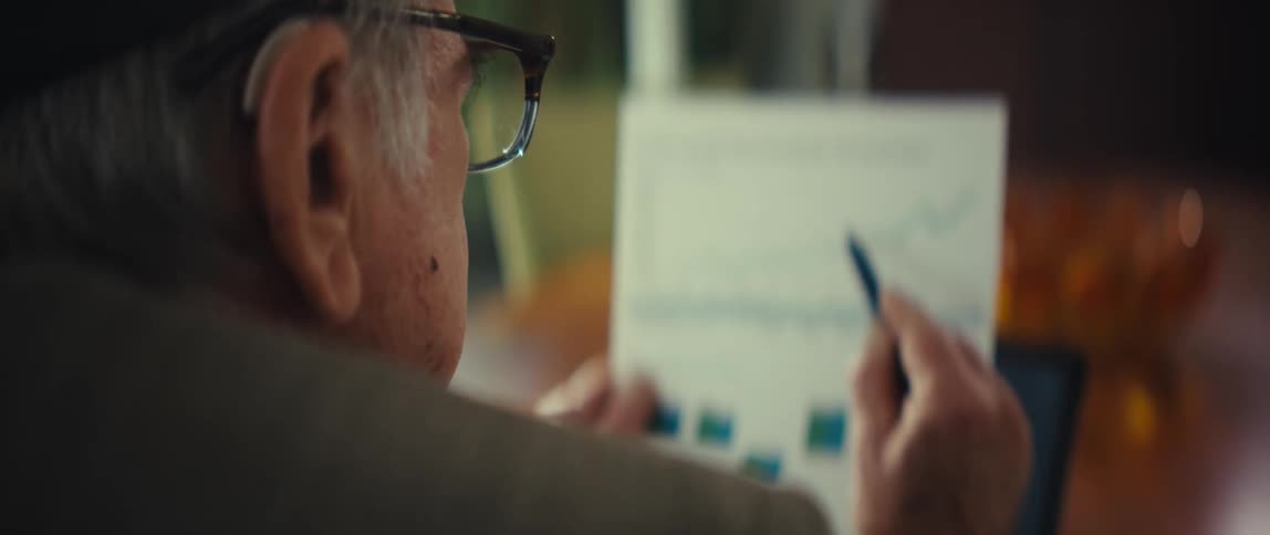 An elderly man in a suit and glasses holds a financial document using a blue pen to track a rising line graph on a portfolio growth chart while sitting at a desk with a monitor in the background