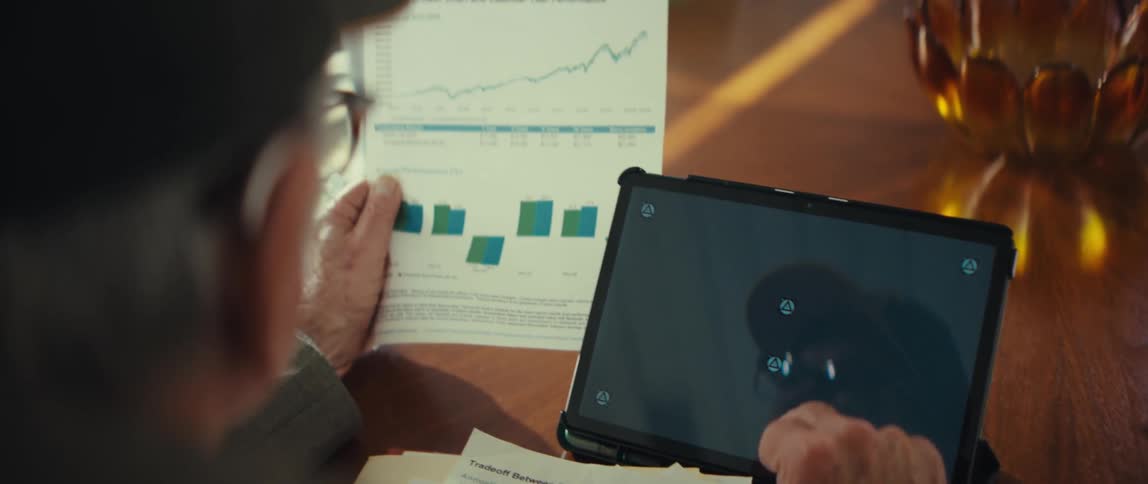 A person reviews financial documents featuring growth charts and performance tables they hold a paper report while simultaneously interacting with a tablet on the table