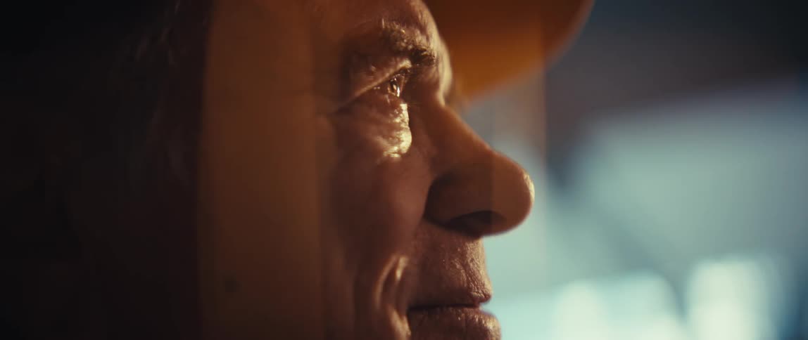 An elderly veteran wearing a black army cap and a grey suit jacket looks toward a window with a peaceful smile as warm sunlight illuminates his face in a bright indoor setting