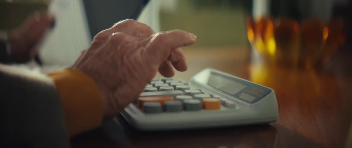 An elderly persons hands are shown in a closeup shot as they press buttons on a large desktop calculatorthe scene captures the deliberate movements of the fingers against the grey and orange keys in a dimly lit indoor setting