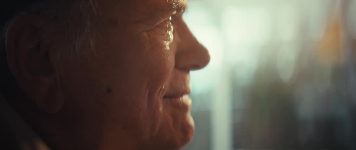 An elderly man sitting indoors wearing a hat with eyes filled with sadness