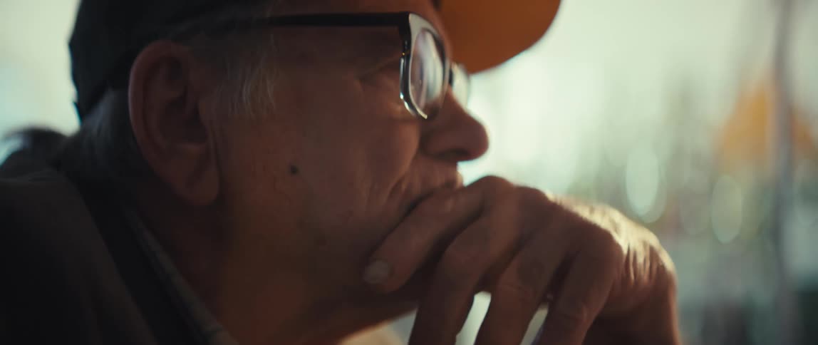 A close view in profile of an elderly man wearing a black cap and glasses as he rests his chin on his hand while looking toward a window with a serious expression in a bright room
