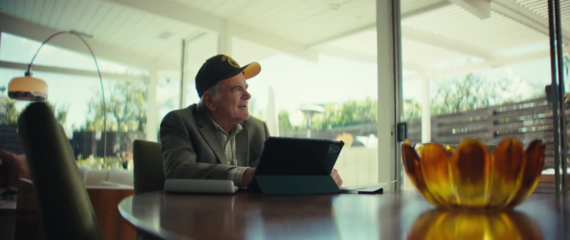 An elderly veteran sits at a dining table with a tablet and paperwork removing his glasses to look away thoughtfully