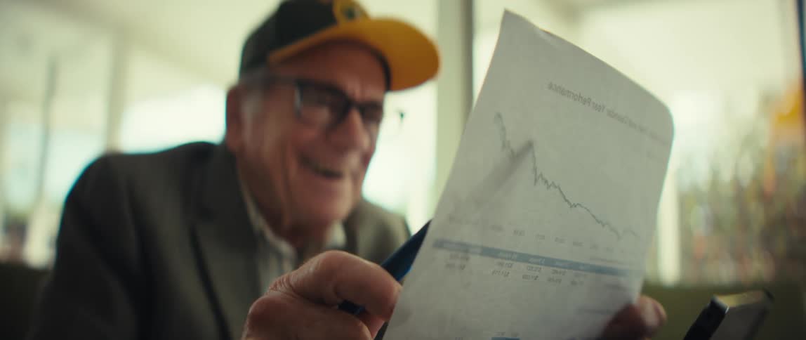 An elderly veteran wearing a black and yellow army cap and a grey suit jacket laughs with joy while looking at a paper with financial charts in a bright room