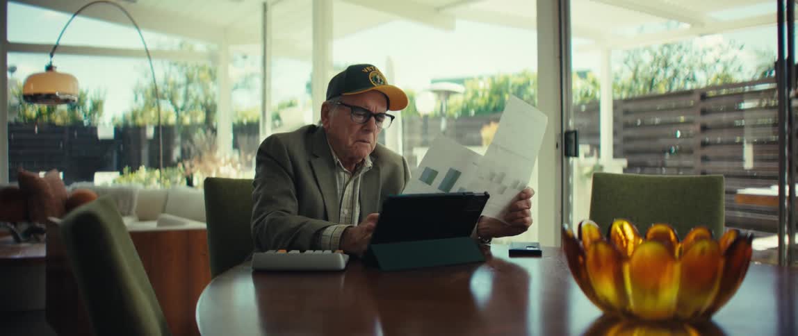 Elderly man wearing a cap and glasses sitting at a table holding a report while looking at a tablet in a room with glass doors in the background