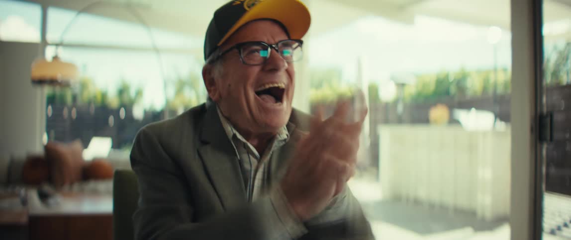 An elderly man wearing a veterans hat and glasses sits at a table looking at documents and reacts with surprise and happiness