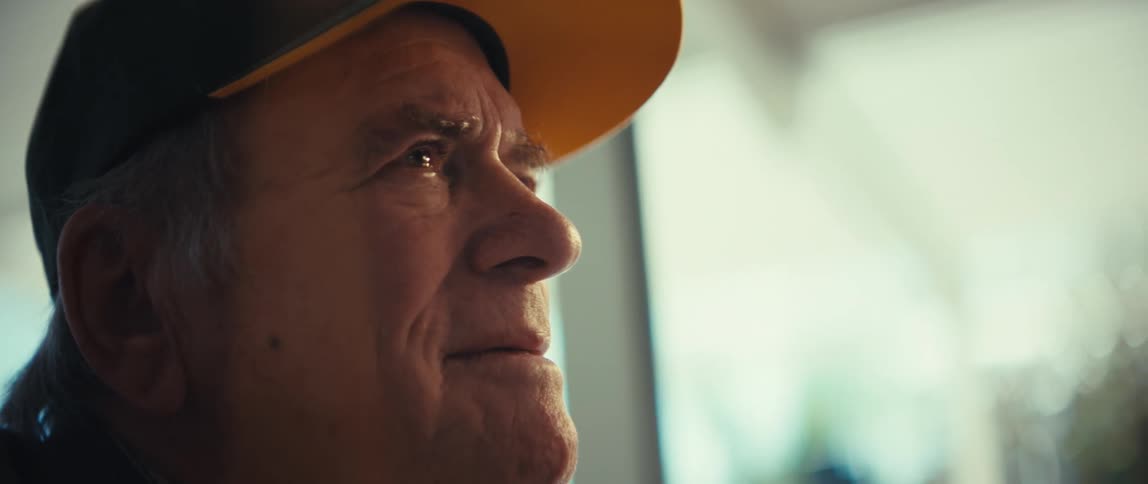 An elderly man wearing a black and yellow baseball cap looks off into the distance with a gentle smile