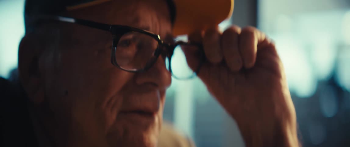 An elderly veteran wearing a black army cap and a grey suit jacket puts on his glasses and rests his chin on his hand in a bright living room