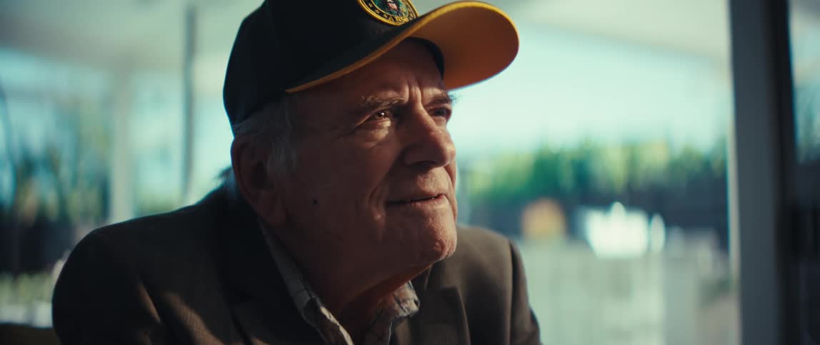 An elderly veteran wearing a black army cap and a gray blazer looks upward with a gentle smile as bright light from a window illuminates his face in a quiet indoor setting