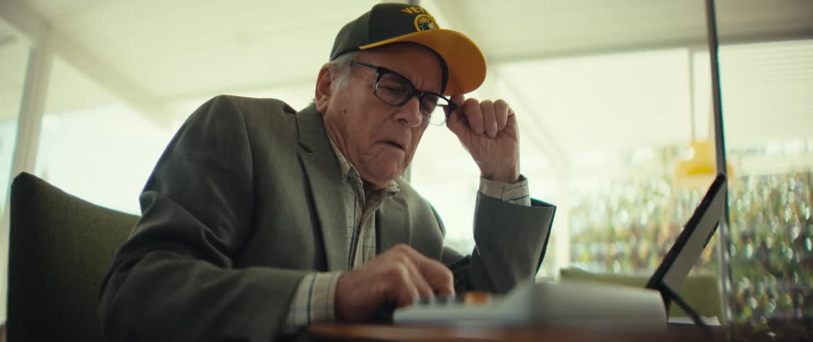 Elderly white man wearing a blazer and a baseball cap sitting in front of a tablet computer using a calculator with a nervous facial expression and then extremely sad indoors during day time