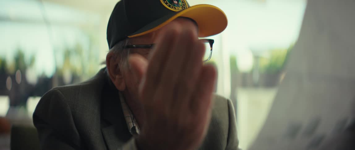 An elderly man wearing a suit and a hat that says veteran puts on his glasses and begins smiling as he reads a document