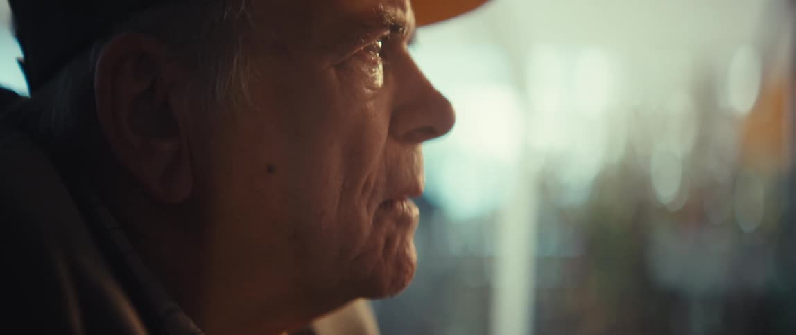An elderly man in a baseball cap looks out of a window with a serious and pensive expression while warm natural light illuminates his face