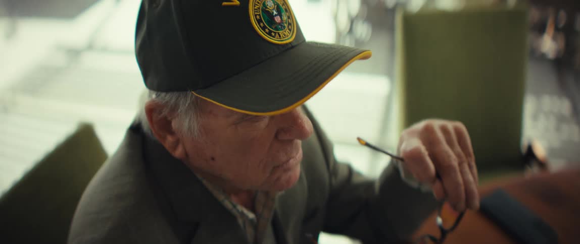 An elderly veteran wearing a black army cap and a grey suit jacket holds his glasses and looks at financial papers before putting the glasses on to read the documents at a wooden table