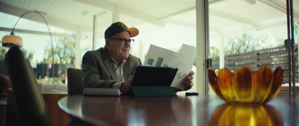 Elderly man wearing a cap and glasses sitting at a table smiling while holding reports in front of a tablet in a room with glass doors in the background