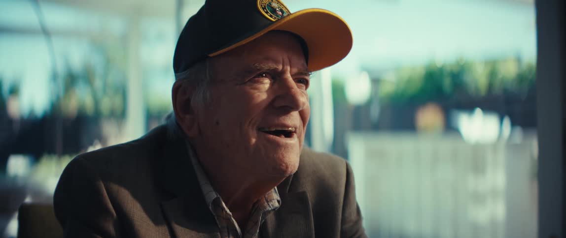 A senior man wearing a brown blazer and a black veteran hat with a yellow brim looks upward and smiles while sitting indoors with a bright blurred background visible through a window