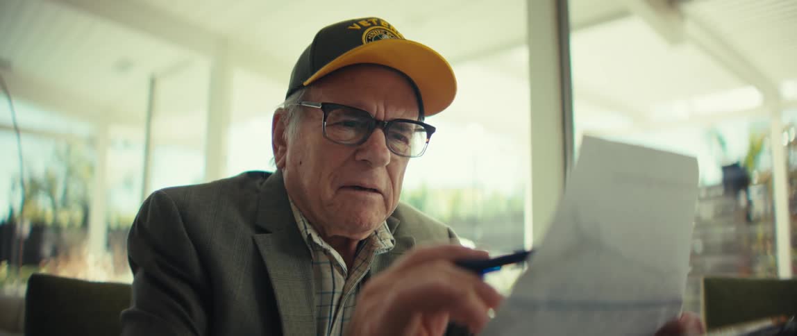 An elderly veteran wearing a black army cap and glasses sits indoors while reviewing a document with a line graph and then looking down with a serious expression