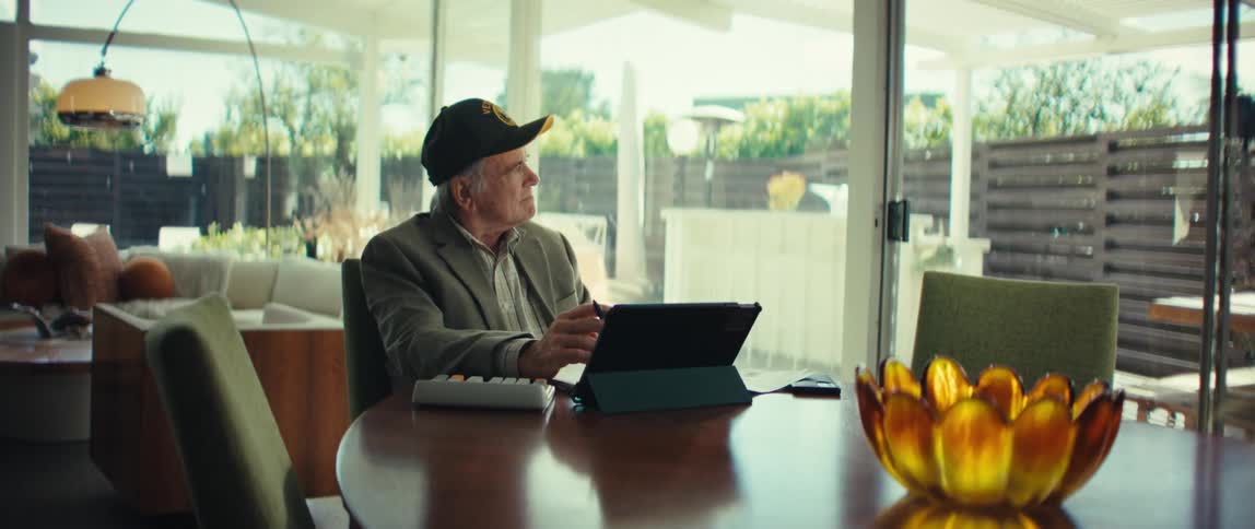 Elderly man wearing a cap sitting at a table with a tablet and calculator while looking through the glass door