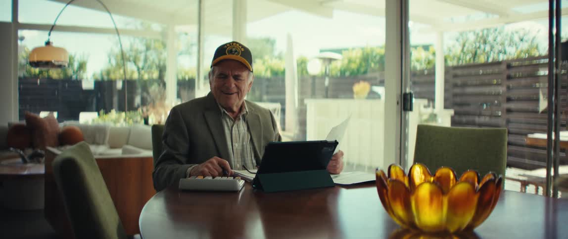 Elderly man wearing a cap sitting at a table using a calculator and a tablet in a room with glass doors in the background