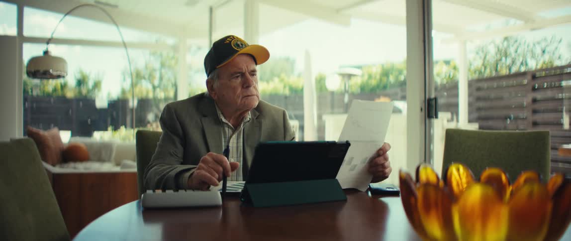 An elderly veteran wearing a black army cap and a blazer sits at a wooden table in a bright room while reviewing bar charts on paper and using a tablet and a calculator