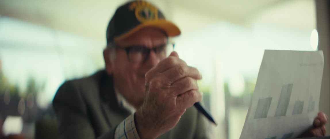 Elderly man wearing a cap and blazer laughing while holding a sheet of paper in a welllit indoor setting